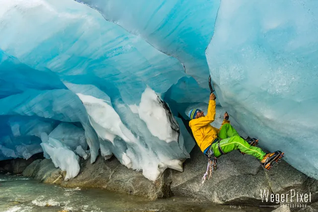Kletterer in gelber Jacke in einer blauen Gletscherhöhle über einem Gletscherbach