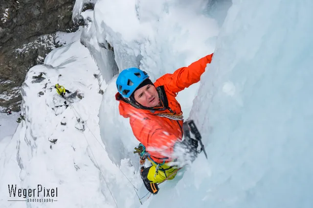 Lachender Eiskletterer mit blauem Helm schlägt die Eisgeräte ins Eis, Eissplitter fliegen