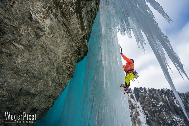 Eiskletterer in orangefarbener Jacke steigt einen türkisblauen Eisfall unter einem Felsdach in Osttirol hinauf