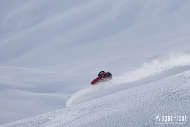 Wintersportler in roter Jacke zieht eine Pulverschneefahne durch einen flachen Hang