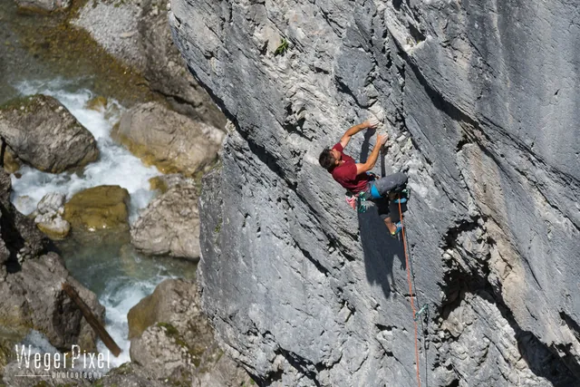 Kletterer in roter Jacke in einer grauen Felswand über einem Wildbach in einer Klamm