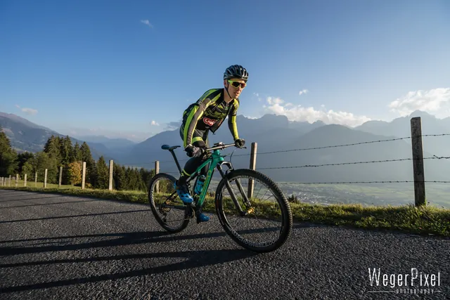 Mountainbiker in grünem Trikot fährt auf einer Bergstraße, dahinter Bergpanorama