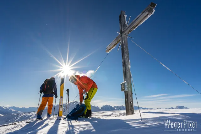 Zwei Skitourengeher am Gipfelkreuz im Gegenlicht der Sonne